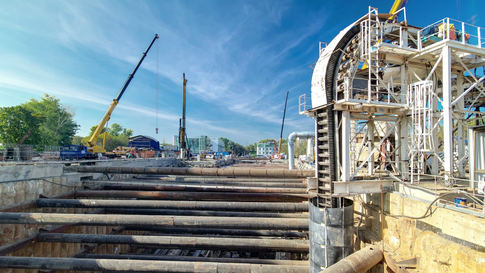 Cranes operating over a large excavation pit at an underground metro station construction site, showcasing heavy-duty beams and facilities robotics in action.